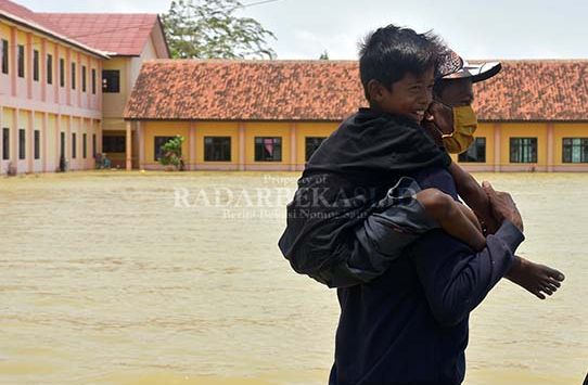 GENDONG ANAK : Seorang anak digendong orangtuanya saat melintasi Sekolah MAN 1 Sukakarya, yang masih digenangi banjir, Senin (22/2/2021). Pemerintah Kabupaten Bekasi belum menangani sekolah-sekollah yang terdampak banjir di wilayah Kabupaten Bekasi.(foto :ARIESANT/RADAR BEKASI)